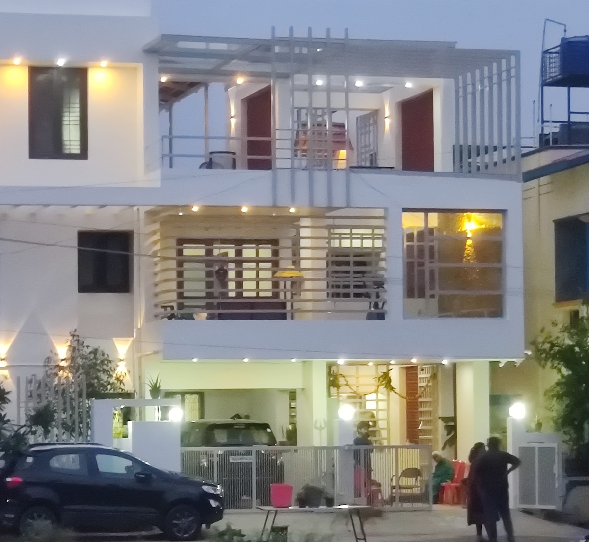 The villa's exterior is shown, featuring a modern design with multiple levels. A spacious balcony is visible, complemented by wooden railing and warm lighting. The ground floor displays large windows and a welcoming entrance. A parked car is seen near the entrance, adding to the accessibility.