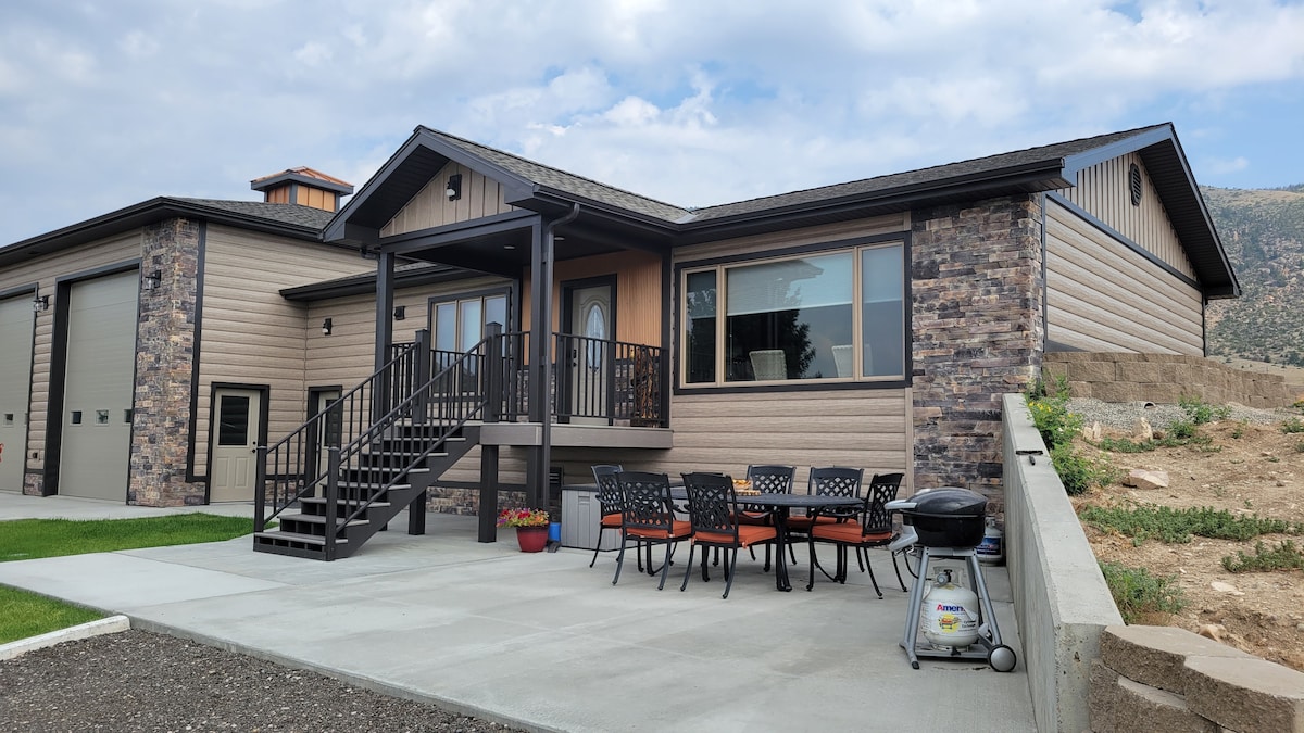 A new mountain home features a welcoming front porch with steps leading to the entrance. A patio area is visible, furnished with a dining table and chairs, surrounded by neatly maintained landscaping. The exterior showcases a mix of siding textures and stone accents, under a cloudy sky.