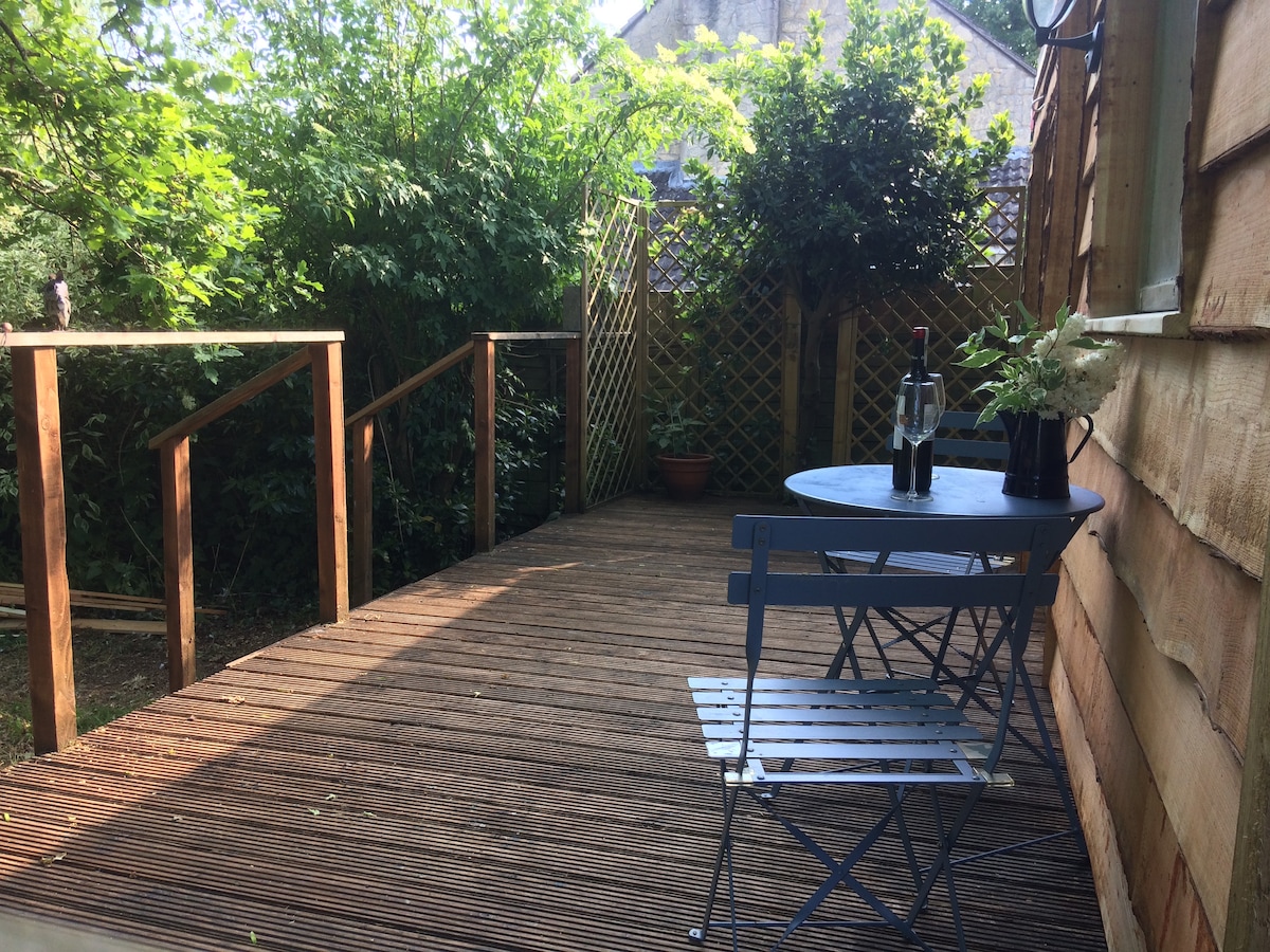 A wooden deck area is surrounded by lush greenery, featuring a small round table with two blue chairs. A bottle of wine and a small vase with flowers are placed on the table. The space is well-lit, creating an inviting outdoor setting.