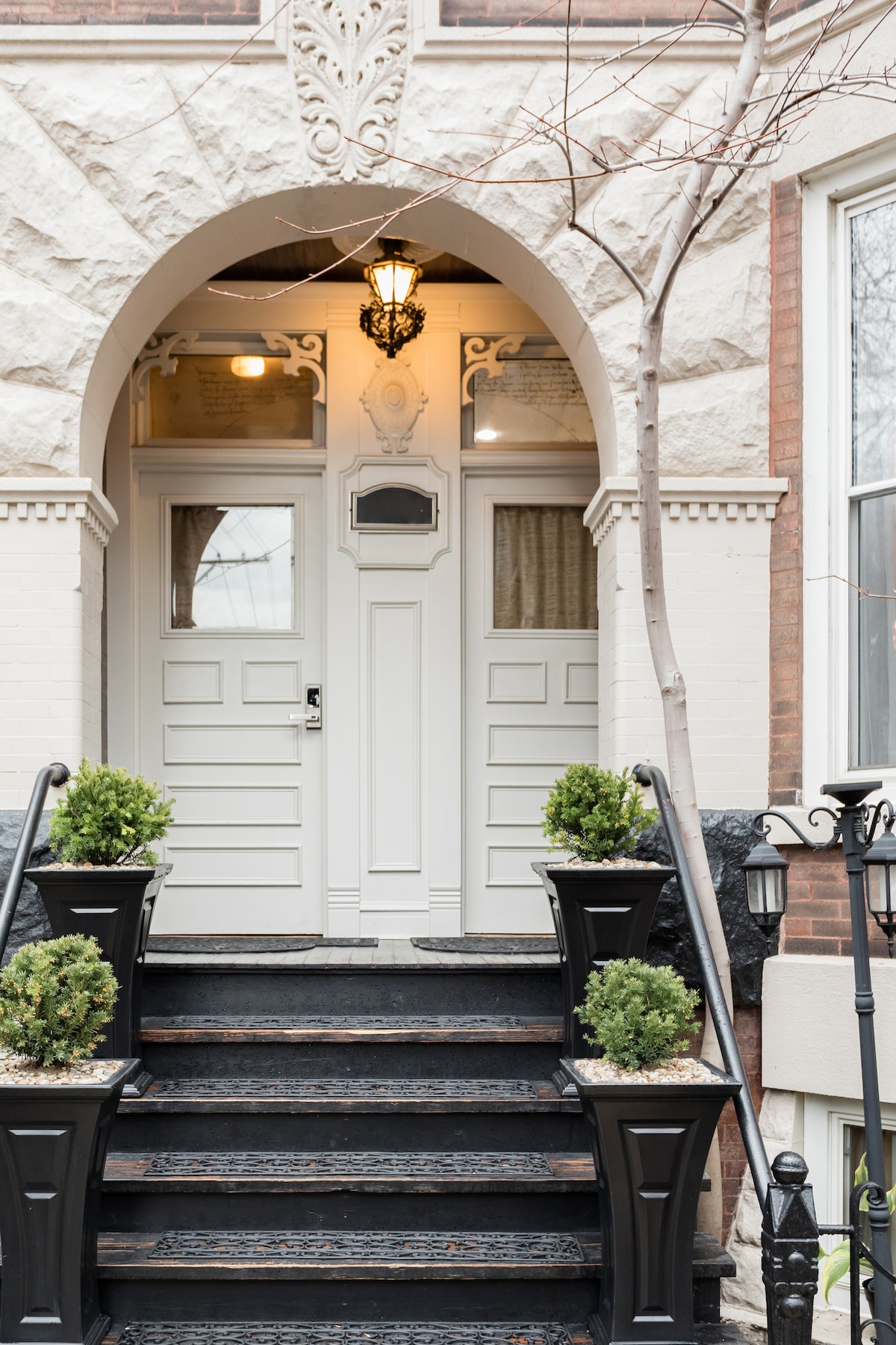 An inviting entrance is marked by a grand archway and elegant light fixtures. Potted greenery flanks both sides of the steps leading up to white double doors, while intricately designed exterior details are showcased on the building's facade.