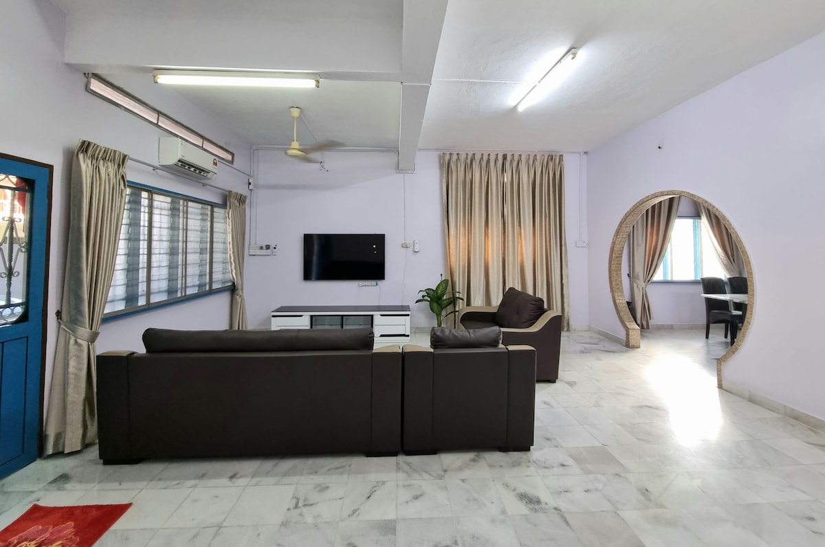 The living room features a spacious layout with comfortable dark sofas facing a wall-mounted television. Soft, neutral curtains frame the large windows, allowing light to enhance the marble flooring. A potted plant adds a touch of greenery, while a curved archway leads to another room.