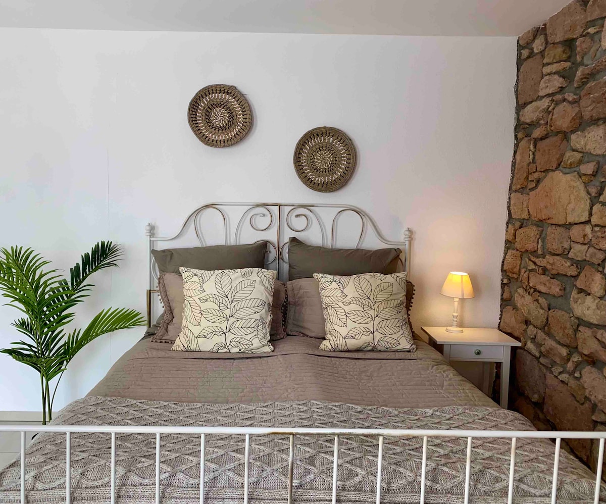 A cozy bedroom features a bed with decorative pillows, set against a textured stone wall. A small bedside lamp provides soft illumination, while a leafy plant adds a touch of greenery to the space.