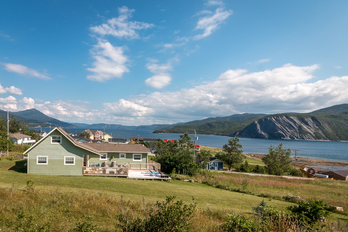 A spacious green chalet is seen from an elevated viewpoint, surrounded by vibrant grassy areas. The coastline and mountains are visible in the background under a clear blue sky, creating a serene backdrop for the property. A wrap-around deck is prominently featured.