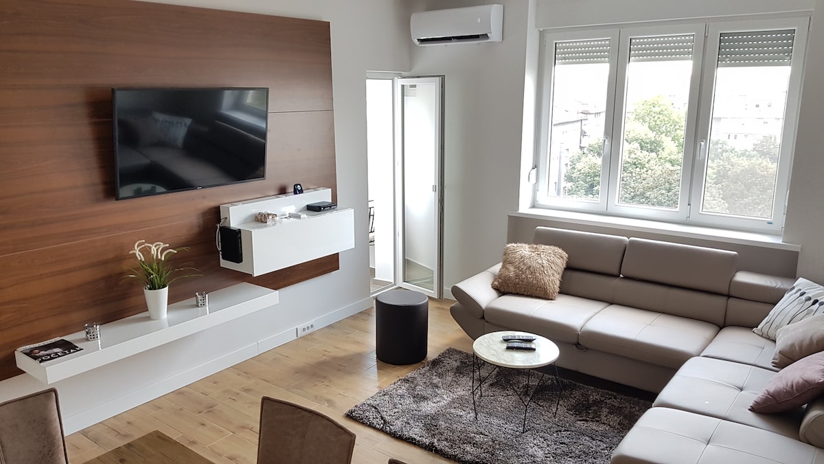 A spacious living room features a comfortable sectional sofa and a round coffee table on a soft area rug. A wall-mounted television is positioned above a modern white shelf. Natural light enters through multiple large windows, and a door leads to another room.