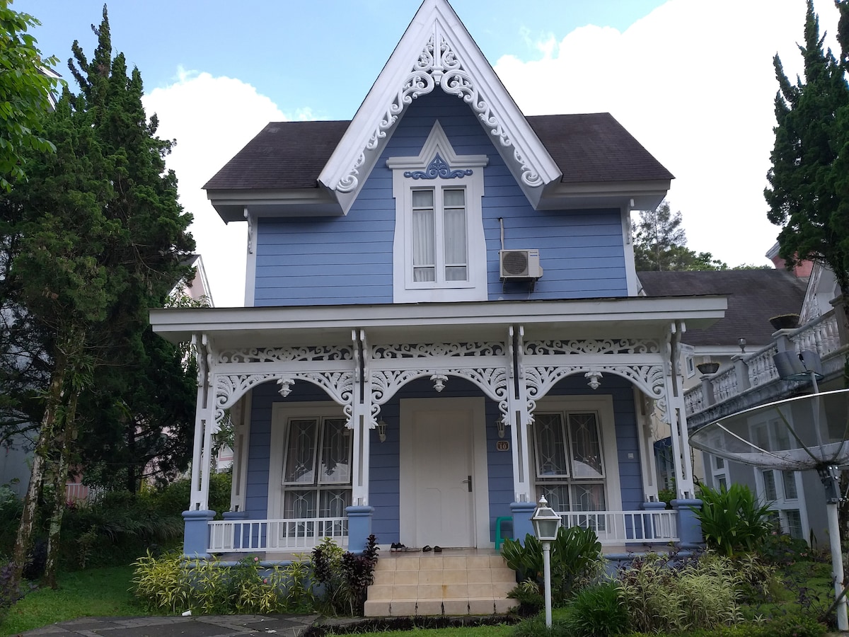 The exterior of the villa is characterized by blue wooden siding and intricate white trim around the windows and porch. Two large windows are framed symmetrically on the upper floor. A well-maintained garden surrounds the entrance, enhancing the home's welcoming appearance.