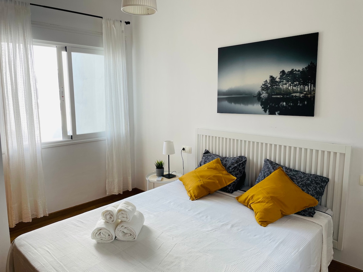 A bright bedroom features a spacious bed adorned with white sheets and yellow accent pillows. A vivid landscape artwork hangs above the headboard. Natural light filters through sheer curtains, illuminating the room. A bedside table with a lamp and a small plant adds a welcoming touch.