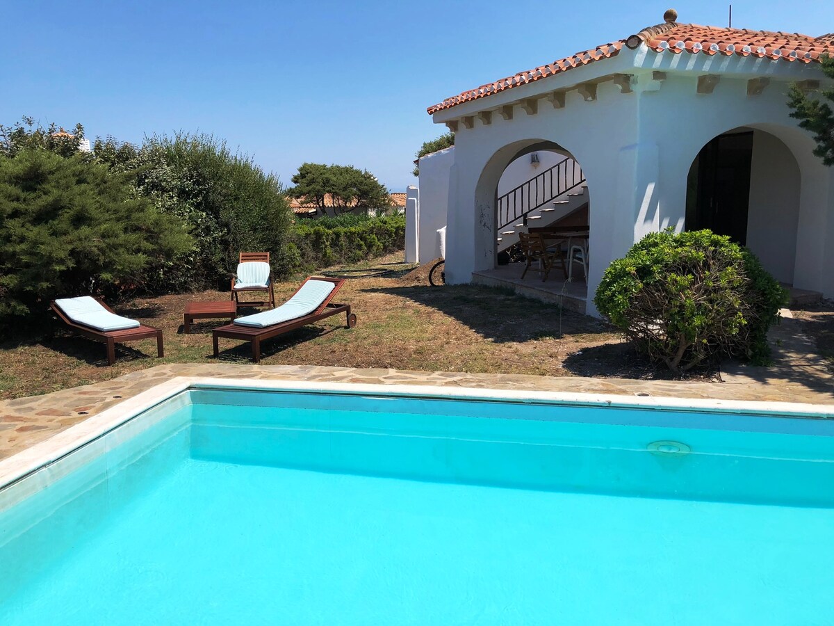 A private pool is framed by grassy areas and a stone pathway. Two lounge chairs are positioned beside the pool, while the exterior of a white villa with a terracotta roof is visible in the background. Bushes and small trees add greenery to the setting.