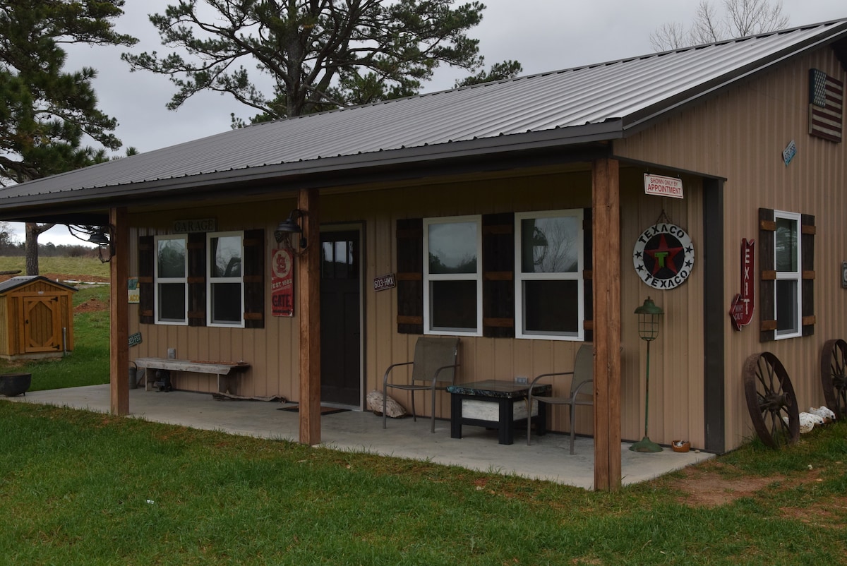 The exterior of the shouse is shown, featuring a covered porch with two chairs and a small table. Large windows allow for ample natural light, while rustic signs and decor adorn the wooden facade. The grassy area surrounding the home adds to its charm.