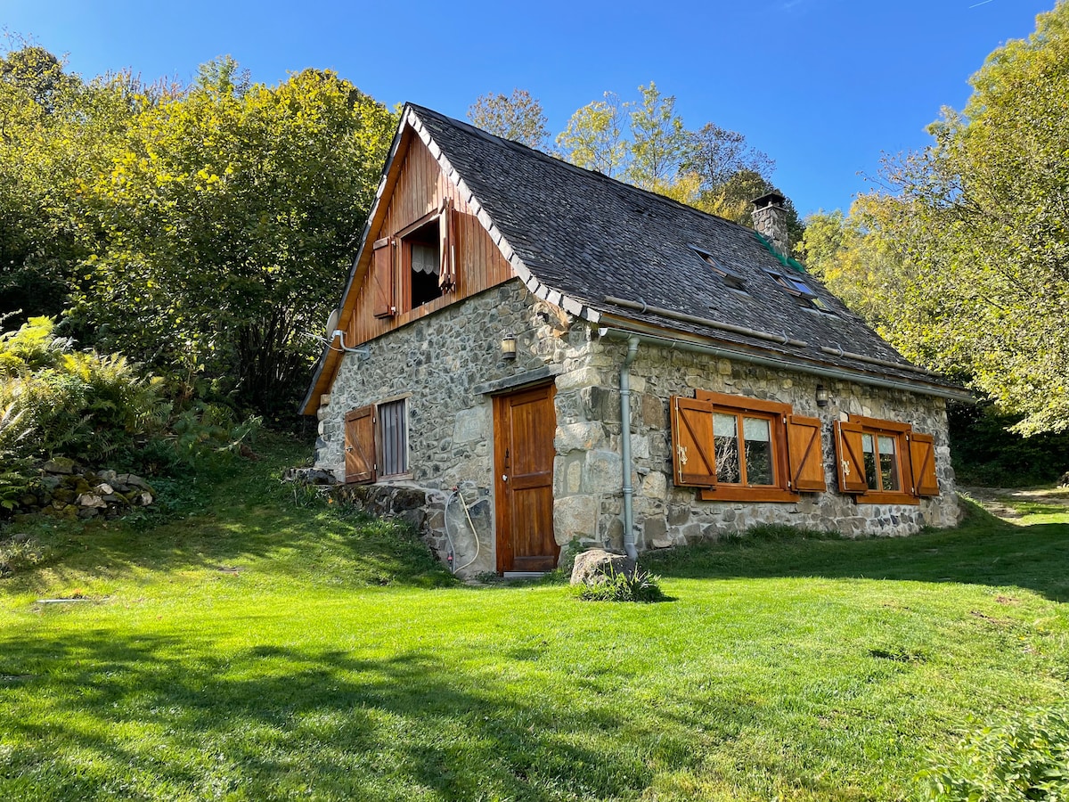 An old stone cottage features a distinctive pitched roof with wooden shutters. Surrounded by green grass and trees, the rustic exterior showcases a natural setting. Warm sunlight highlights the textures of the stone, creating a serene atmosphere for a getaway.