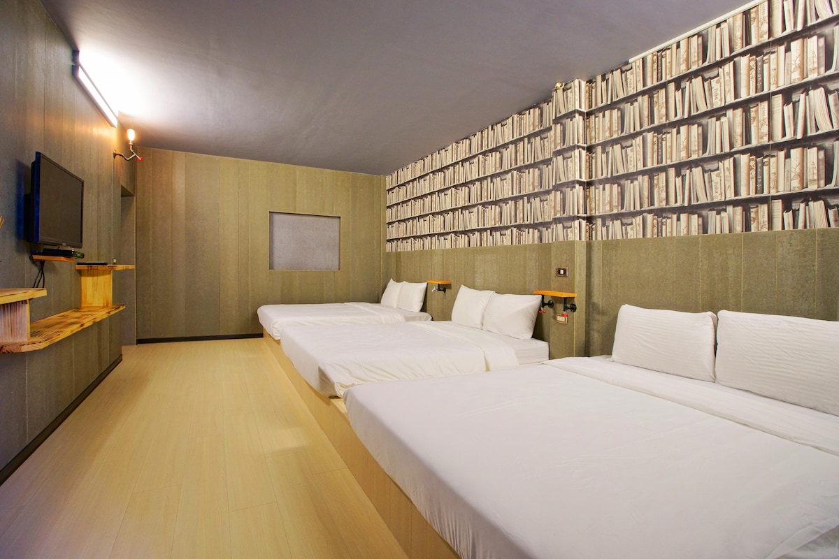 A spacious guest room is furnished with three beds, each made with white linens. A feature wall is decorated with a pattern resembling stacked books, and a television is mounted on the wall opposite the beds.