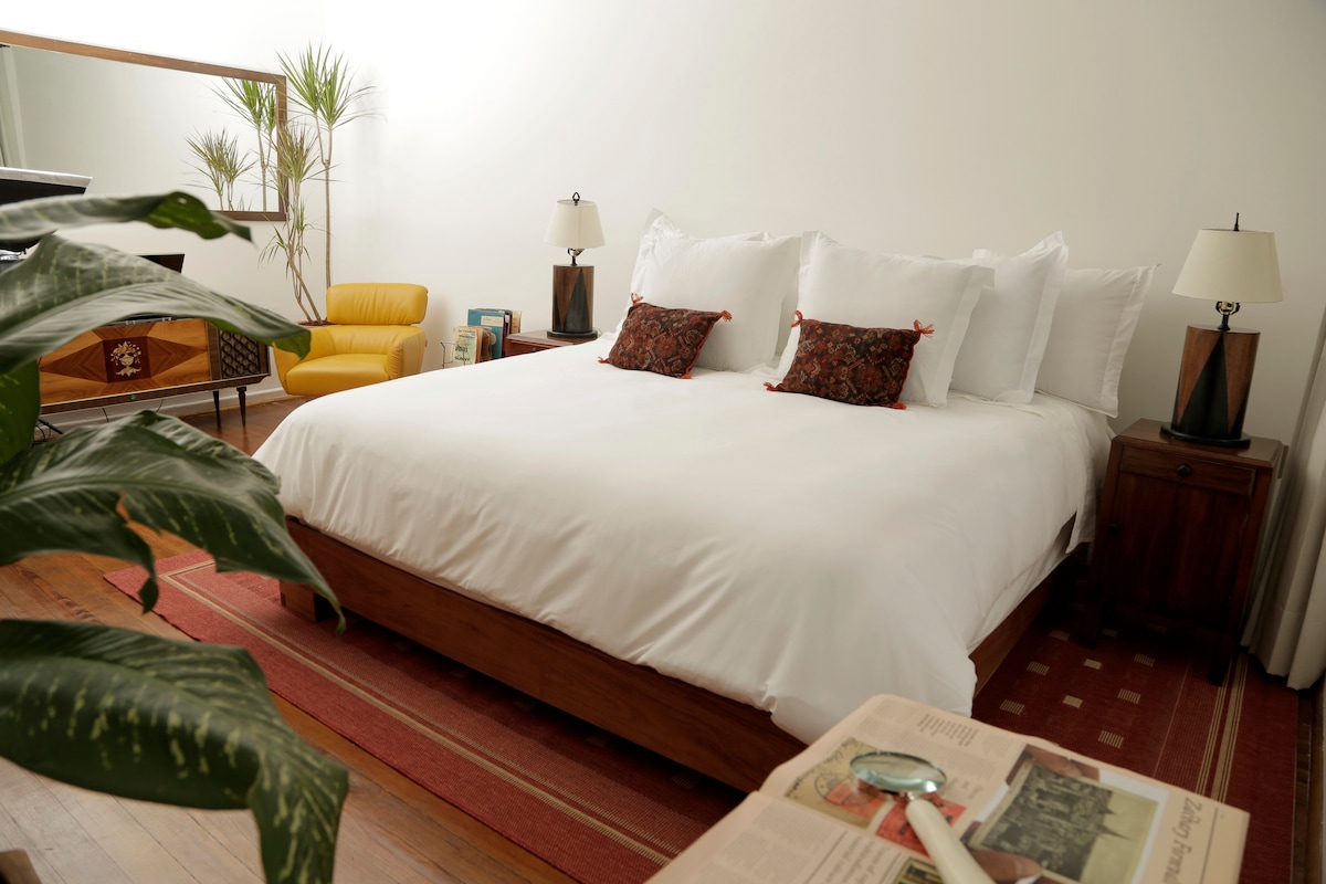 A spacious sleeping area features a large bed adorned with white linens and decorative pillows. Simple nightstands with lamps flank the bed. A cozy yellow chair and a potted plant add warmth, while a wooden dresser and a coffee table with a newspaper provide functional elements.