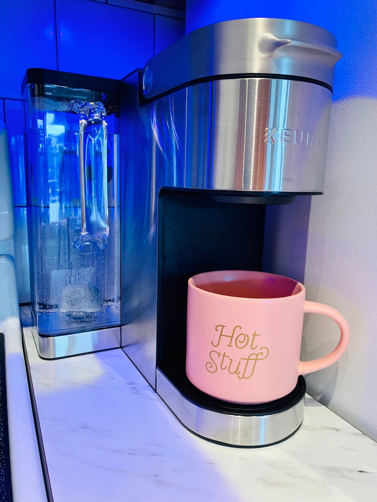 A Keurig coffee maker is positioned on a marble countertop, beside a water reservoir. A pink mug with the inscription 'Hot Stuff' sits on the drip tray, ready for brewing. The area is subtly illuminated by bright blue kitchen lights.