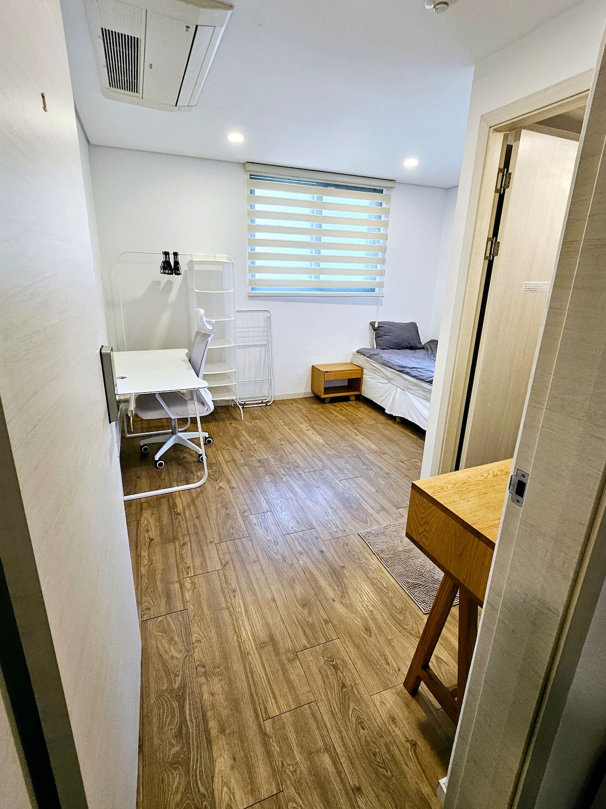 A bright room is captured with wooden flooring and a compact layout. A spacious bed is visible along with a desk and chair, ideal for studying. Natural light filters through the window, and a small wooden nightstand is positioned beside the bed.