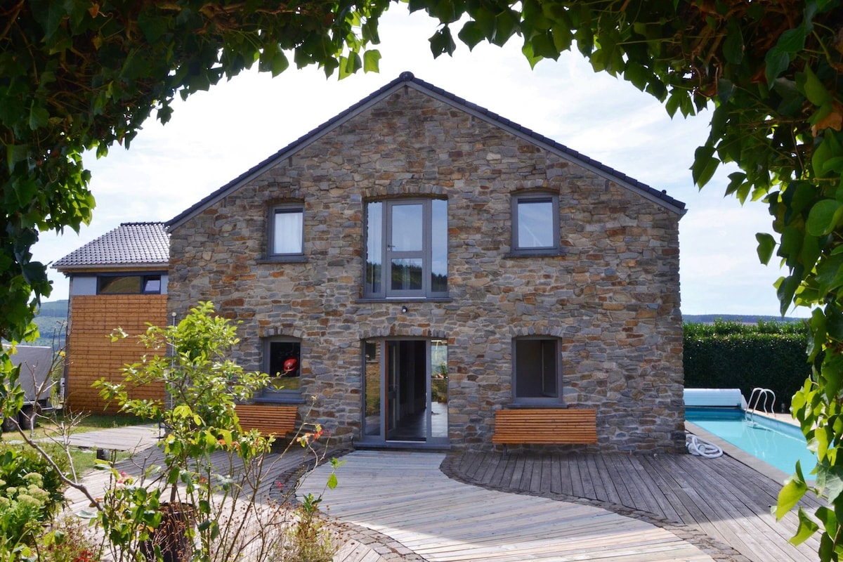 A stone-clad holiday home is framed by greenery, showcasing a wooden deck leading to the entrance. The façade features large windows that brighten the interior. A private swimming pool is visible in the background, inviting relaxation in the garden setting.