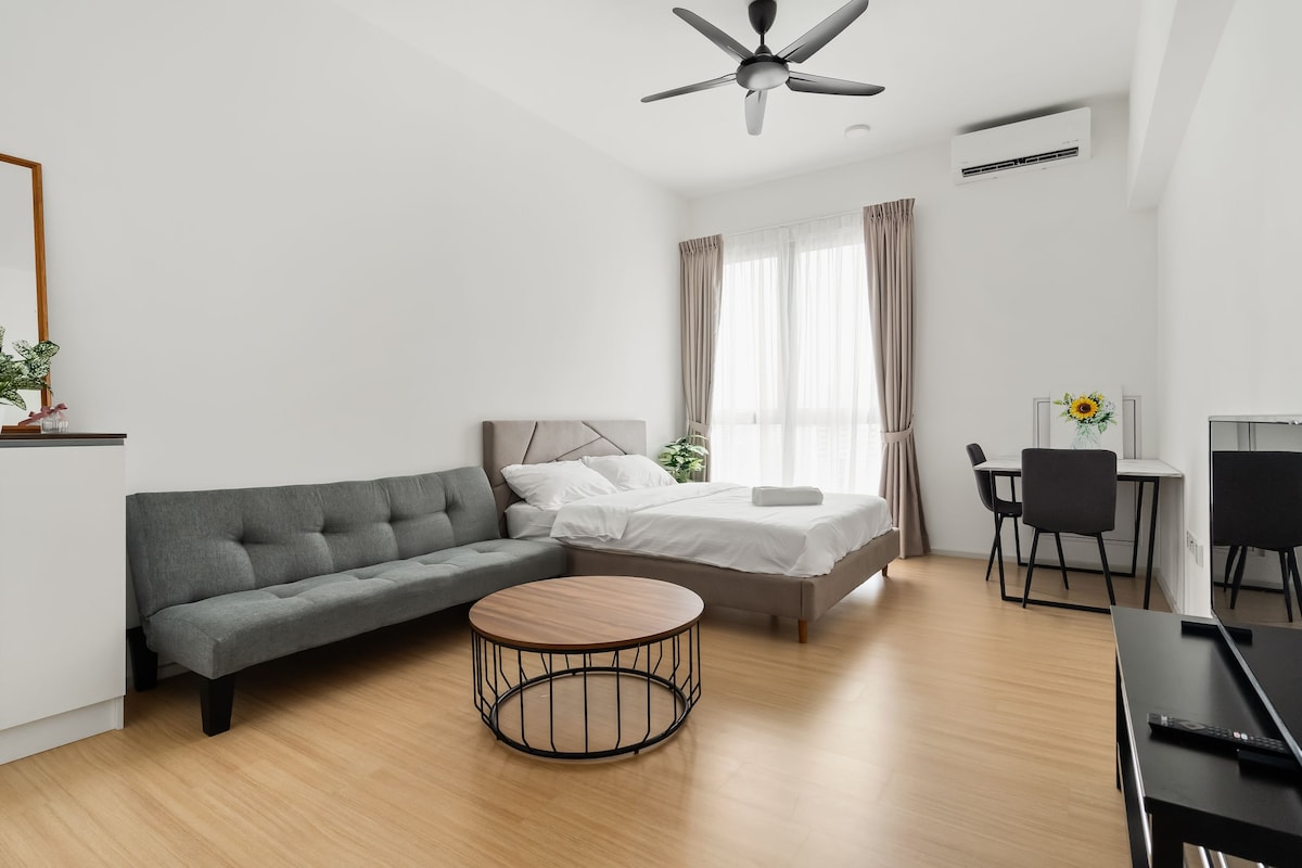 A cozy studio apartment features a queen bed with a light-toned comforter and a gray sofa bed. The room is illuminated by natural light from large windows, draped with sheer curtains. A dining table with chairs and a coffee table are positioned in the welcoming space.