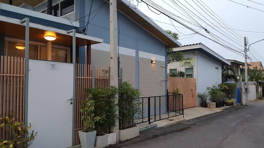 A charming exterior is presented with two light-colored buildings situated side by side, featuring wooden accents and greenery. A wide walkway leads towards the entrance, bordered by planters, while power lines and a street view provide context to the surroundings.