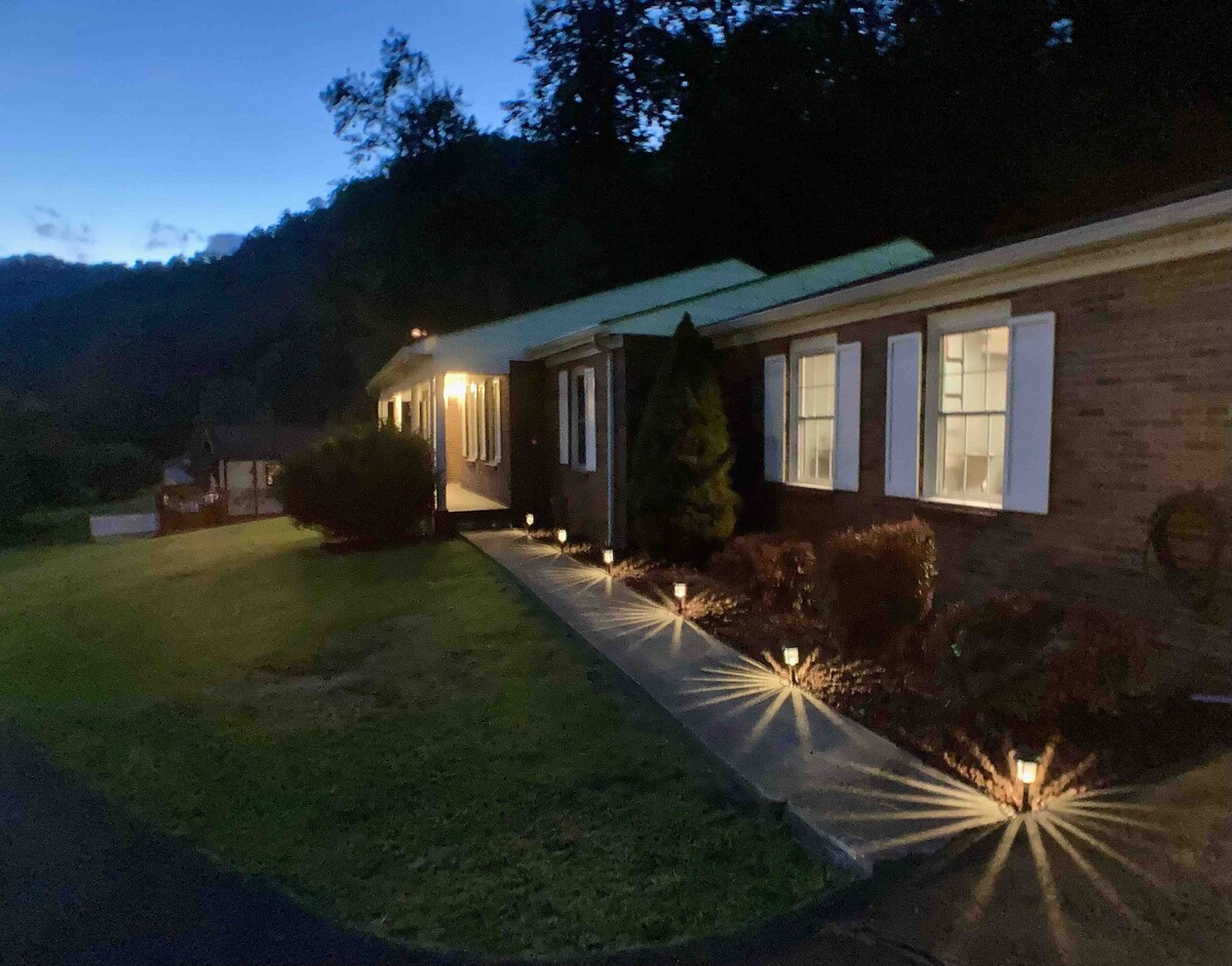 The exterior of a single-story brick home is seen at twilight, with outdoor lighting illuminating the pathway and surrounding landscaping. Flanked by well-maintained hedges, white shutters complement the windows, creating a balanced and inviting facade. The backdrop features gentle hills and a clear evening sky.