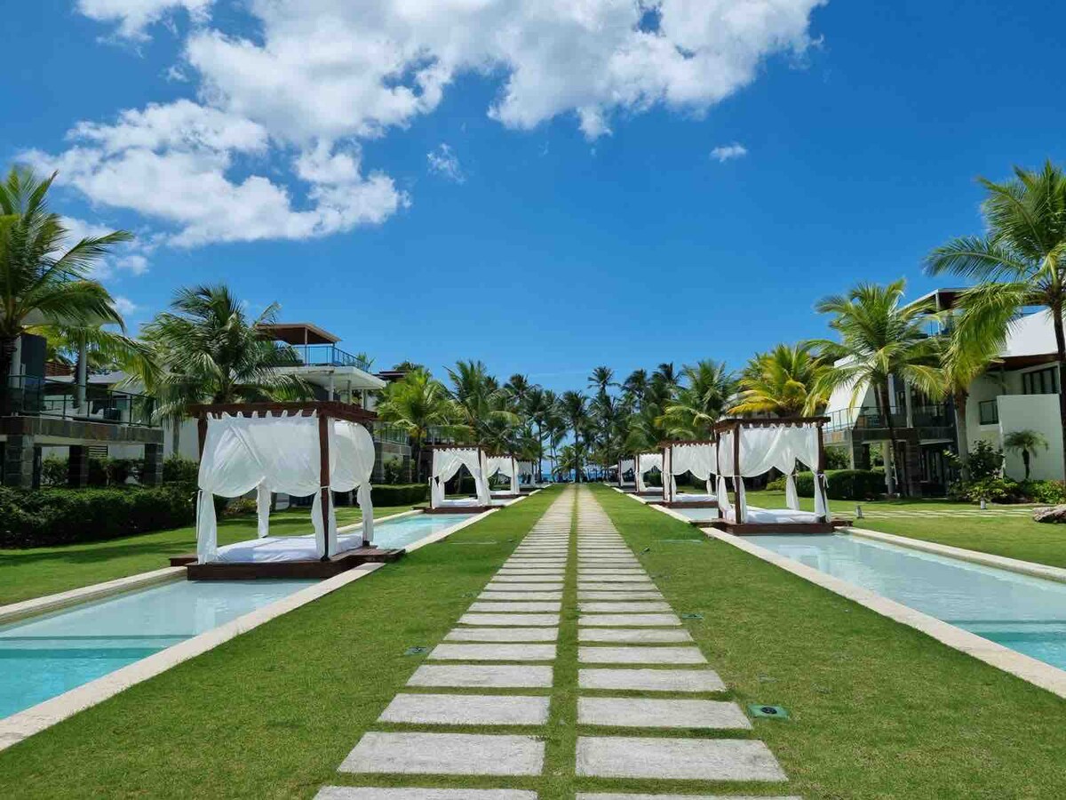 A serene pathway lined with lush greenery leads toward a row of elegant cabanas on either side. Crystal-clear pools run parallel, reflecting the vibrant blue sky and palm trees, creating a tranquil and inviting atmosphere for relaxation.