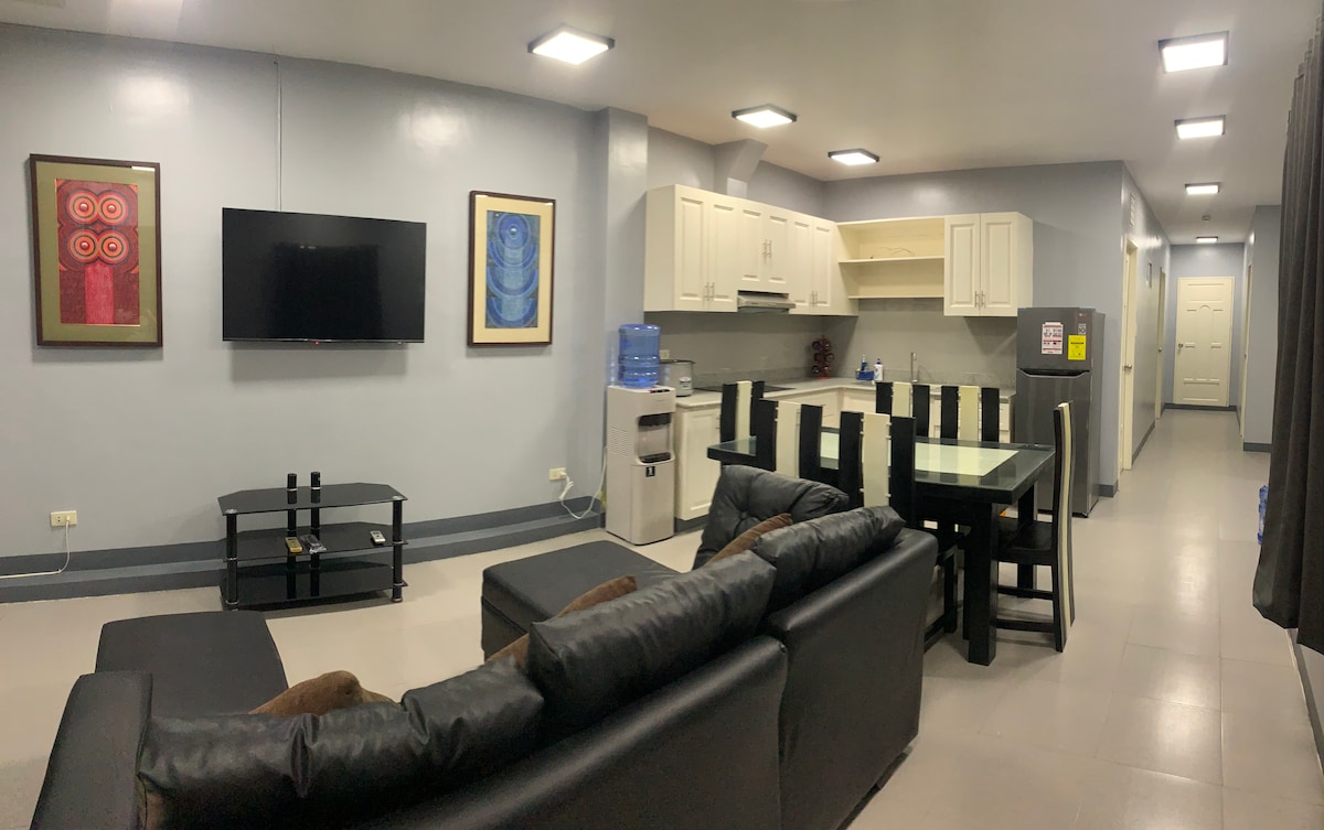 The open living area features a black sectional sofa, a flat-screen television mounted on the wall, and a glass dining table surrounded by black chairs. The modern kitchen is equipped with white cabinets, a refrigerator, and a water dispenser, with light grey walls enhancing the spacious ambiance.