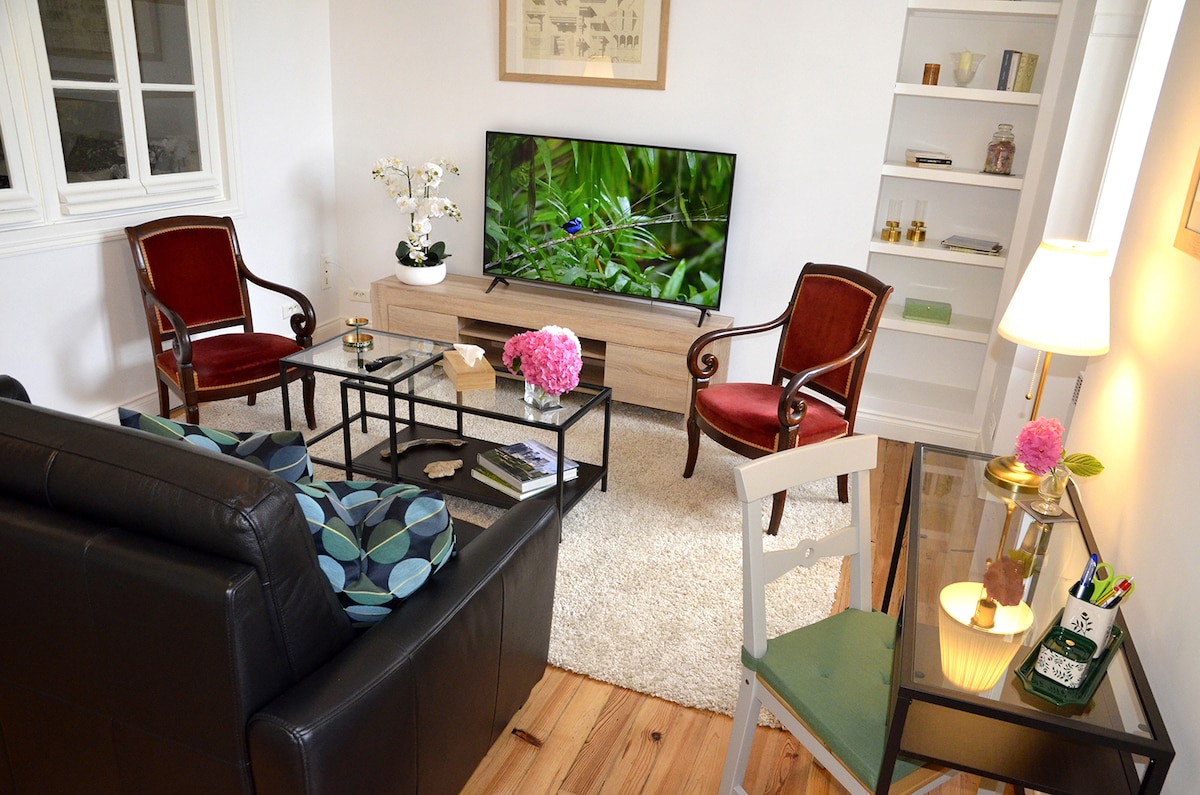 A cozy living area is presented with a large, dark sofa accented by colorful cushions. Two vintage red armchairs flank a low glass coffee table, which sits atop a soft area rug. A flat-screen television offers entertainment, while natural light fills the space through nearby windows.