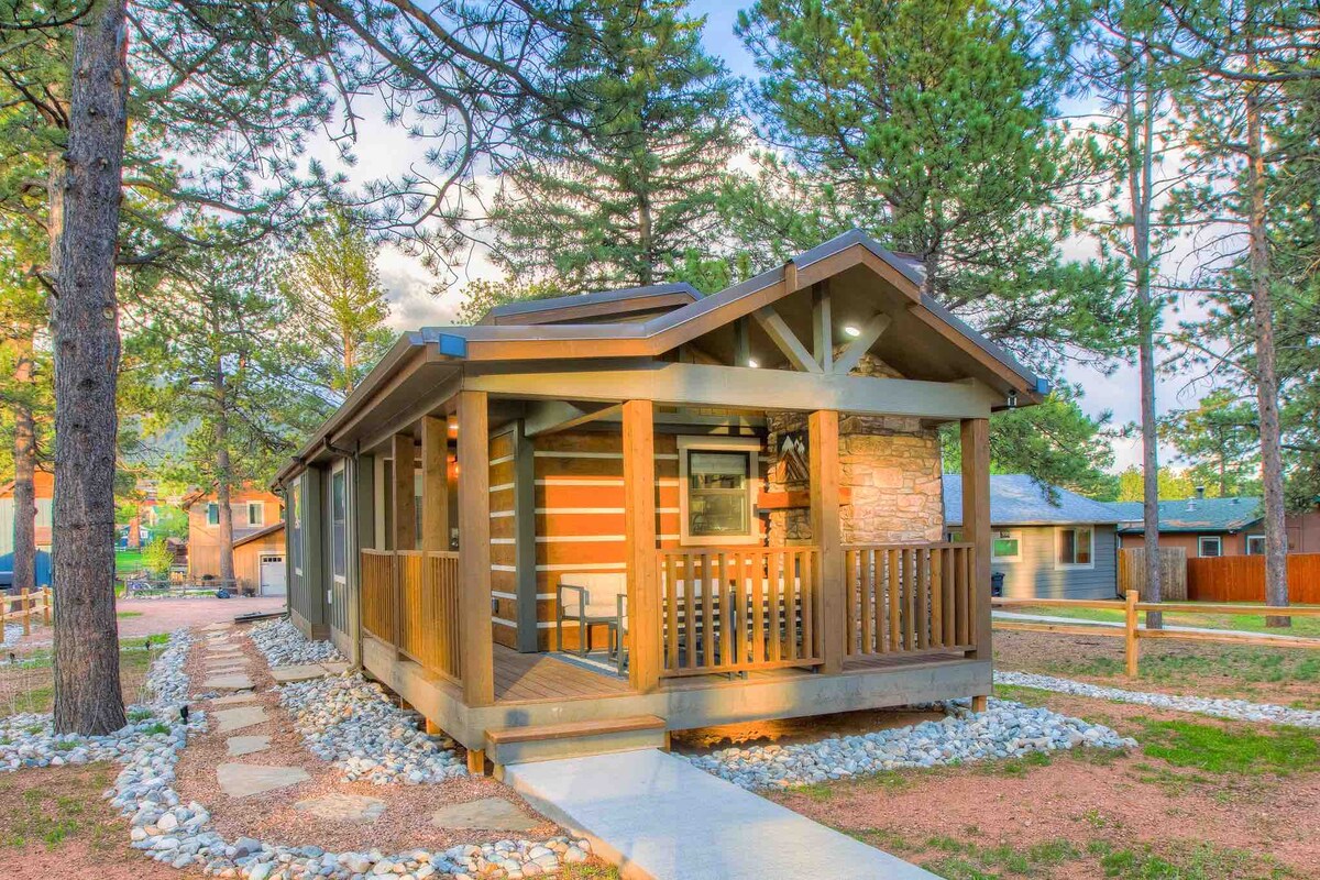 A charming cabin features a wooden porch with a stone accent wall, surrounded by tall pine trees. A well-maintained pathway leads to the entrance, with pebbles lining the walkway. Natural light highlights the rustic design and outdoor seating area.