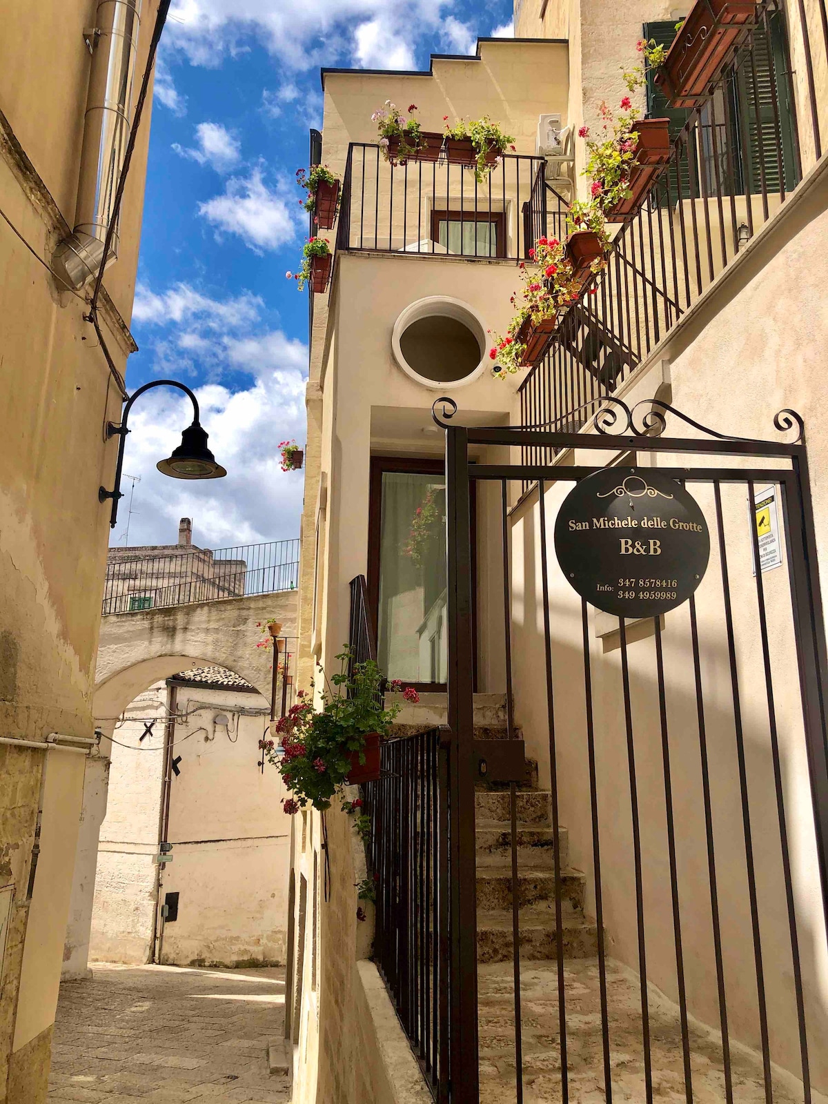 A charming entryway is framed by wrought iron gates adorned with vibrant flower pots. The building's facade features a mix of smooth and textured surfaces, with a glimpse of the surrounding narrow street and nearby architectural elements visible in the background.
