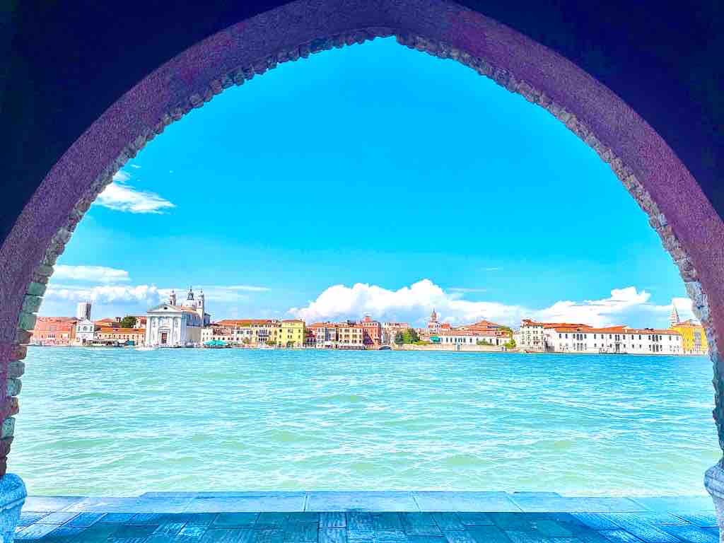 A view through an arched window showcases the vibrant landscape of Venice. Colorful buildings line the waterfront, with clouds reflected on the shimmering water. The scene conveys a sense of tranquility, inviting exploration of the city's historic architecture and scenic waterways.