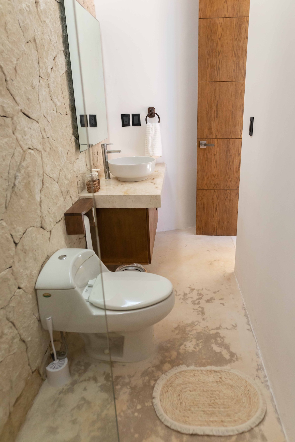 A clean and modern bathroom features a rustic stone wall and a wooden cabinet. A round sink sits atop the countertop. A toilet and a plush rug are positioned on the smooth floor, while soft natural light enhances the serene ambiance.