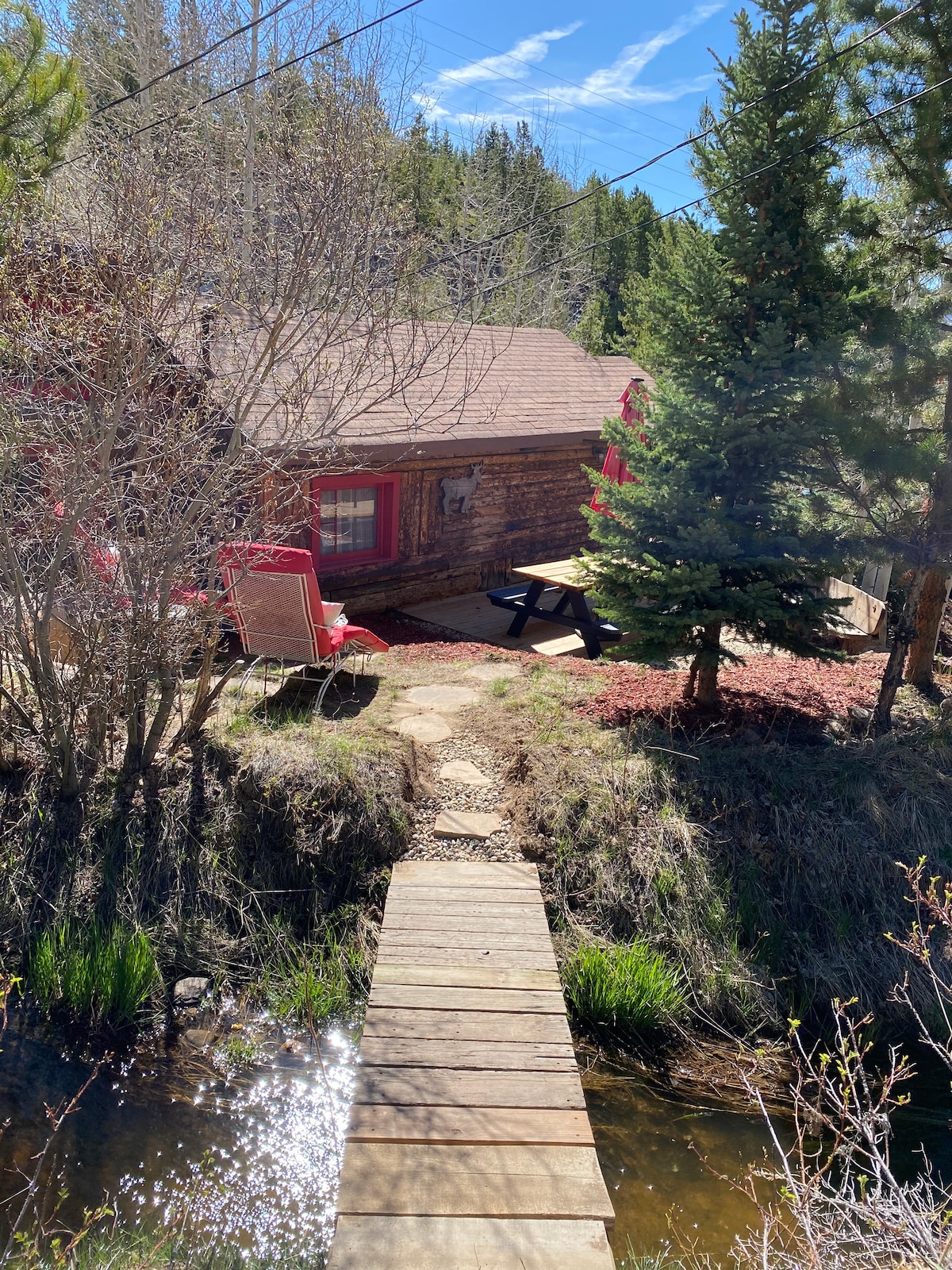 A rustic cabin is nestled among trees, accessible via a wooden bridge over a tranquil stream. Red accents on the cabin’s windows complement the surrounding greenery. A picnic table and two chairs create a cozy outdoor space, perfect for enjoying the natural setting.