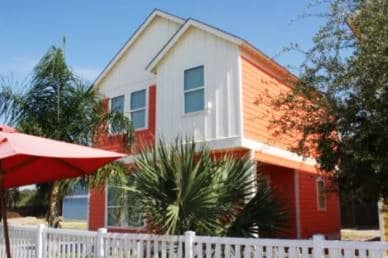 A two-story coastal home features an orange exterior with white accents. Large windows allow natural light to fill the space, surrounded by lush palm trees. A white picket fence provides a charming boundary, with a red umbrella visible in the foreground.