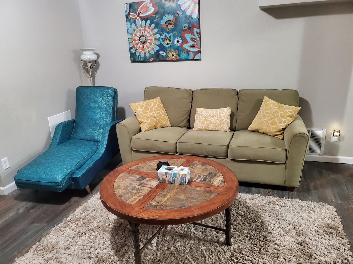 A comfortable seating area is featured, including a green sofa adorned with two patterned cushions and a blue lounge chair. A circular wooden coffee table occupies the center, topped with decorative items. A colorful wall art piece adds a touch of vibrancy to the space.