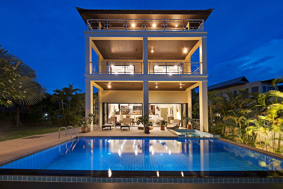 The three-storey villa is showcased at dusk, illuminated by warm exterior lights. A spacious swimming pool reflects the structure, while lush tropical plants surround the area. The open ground floor living space can be seen through large glass doors, enhancing the connection to outdoor living.