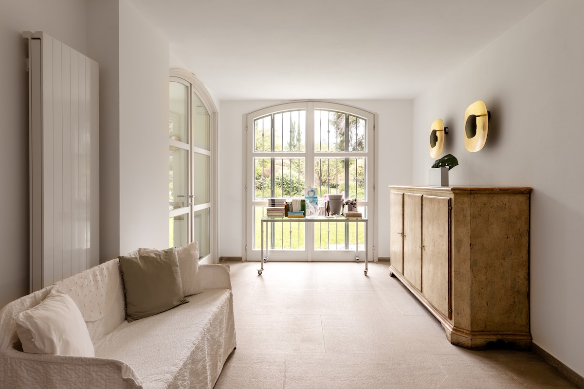 A spacious living area is adorned with a neutral-toned sofa and an ornate sideboard. Large windows allow natural light to fill the room, offering views of the lush garden outside. Soft lighting fixtures create a relaxed ambiance.