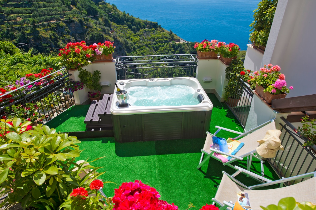 A private terrace features a jacuzzi surrounded by vibrant flowering plants, offering a view of the sea. Two lounge chairs are positioned nearby, along with a small table holding refreshments. The space is designed for relaxation in a serene outdoor atmosphere.