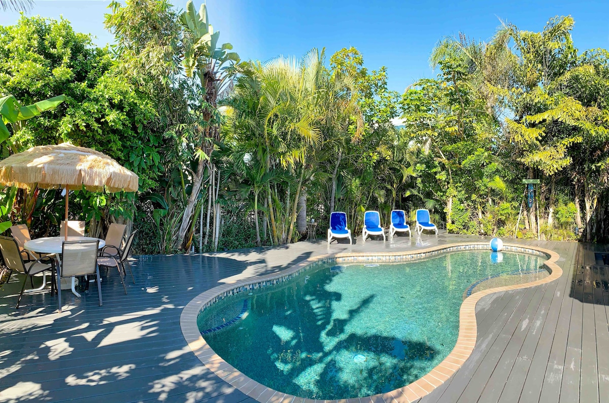 A private heated saltwater pool is set within a lush tropical landscape, surrounded by palm trees and vibrant greenery. Comfortable lounge chairs are positioned nearby, along with a shaded outdoor dining area featuring a thatched umbrella. Sunlight glistens on the water's surface, creating a serene atmosphere.