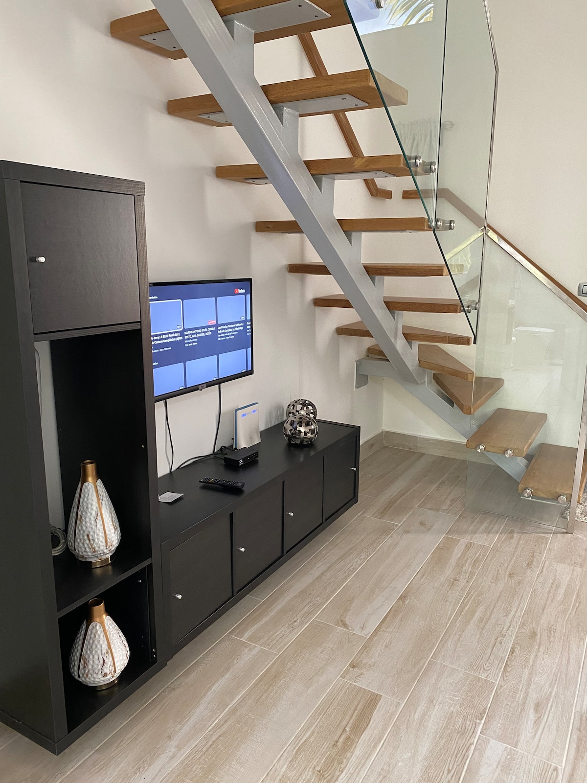 A modern living space features a sleek entertainment unit with decorative vases and a flat-screen television mounted on the wall. A staircase with wooden steps and glass railings leads to the upper level. Neutral-toned tiles cover the floor, creating a bright and spacious environment.