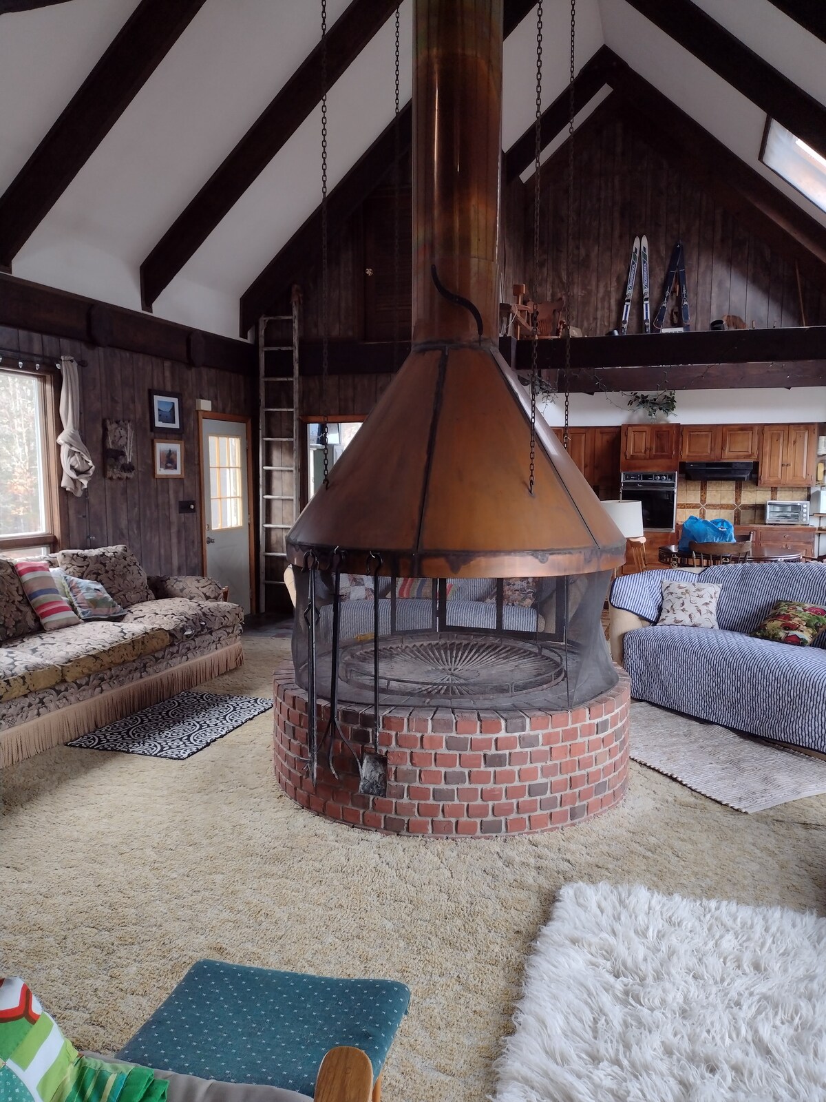 A striking central fireplace made of brick and metal is featured in the living area, surrounded by cozy seating options. The space is adorned with natural wood beams and large windows, allowing for ample light. Textured rugs cover the floor, enhancing comfort.