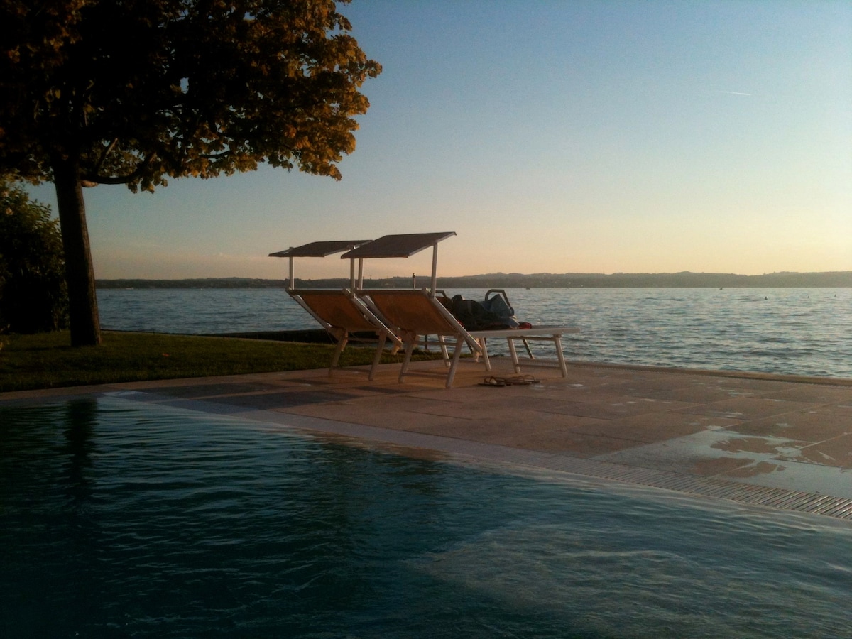 A serene outdoor area features a poolside view of the lake, complemented by two lounge chairs beneath shaded awnings. The setting sun casts a warm glow over the tranquil water, creating a peaceful atmosphere for relaxation.