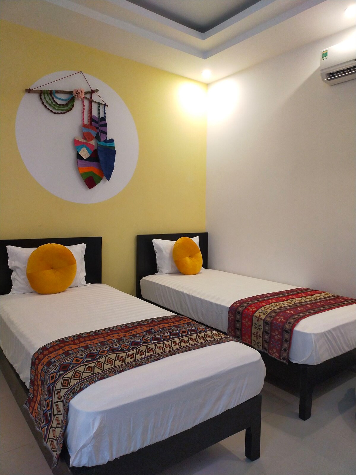 A bright sleeping area features two separate beds, each dressed in crisp white linens. Colorful patterned throws are layered across the beds. A decorative wall hanging consisting of various textiles is displayed above, adding a playful touch to the yellow accent wall.