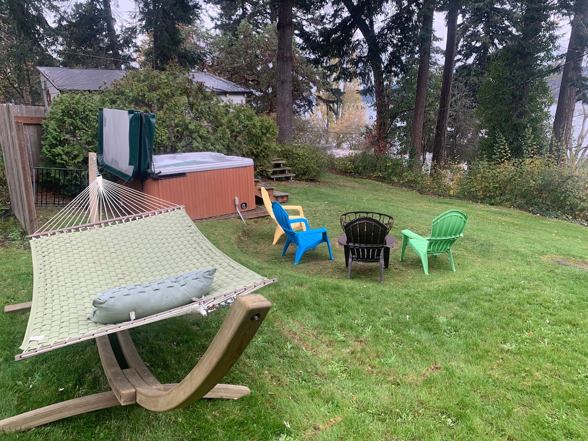 A spacious yard features a green hammock suspended between wooden posts. Nearby, a private hot tub is positioned against a backdrop of trees. Colorful chairs in blue, green, and black are arranged around a small fire pit, creating an inviting space for relaxation.