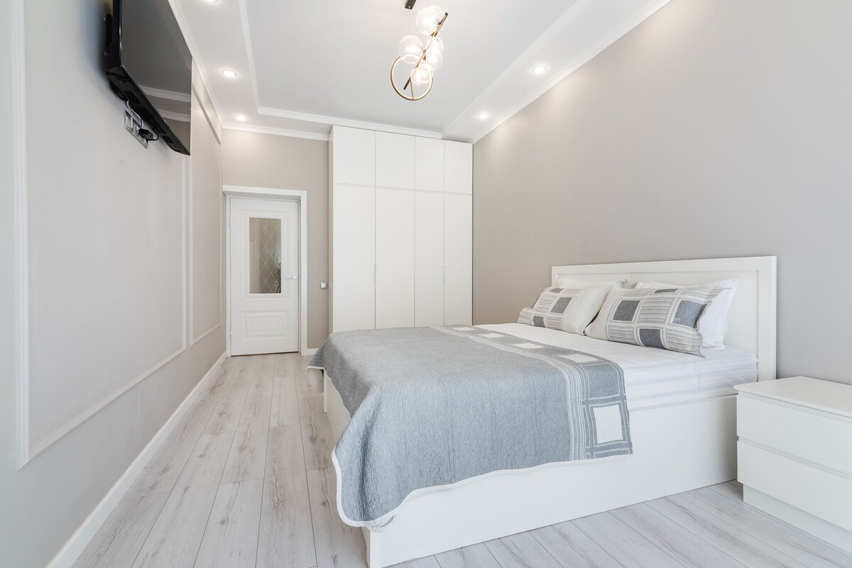 A well-appointed bedroom features a spacious bed with a light grey throw and patterned pillows. Natural light illuminates the room, highlighting the light-colored walls and flooring. A modern light fixture and wall-mounted TV enhance the contemporary design.
