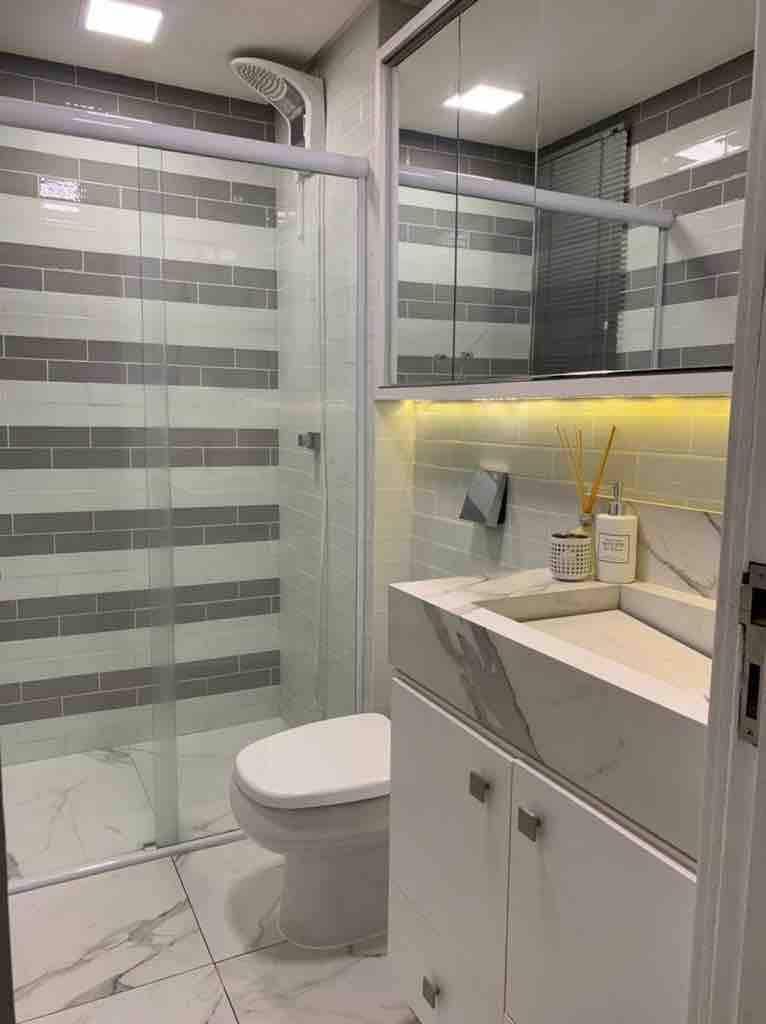 A modern bathroom is presented, featuring a walk-in shower with glass doors and a wall lined with horizontal tiles in varying shades of grey. A white vanity with a marble countertop holds toiletries, complementing the sleek design. Natural light reflects off the glossy surfaces.