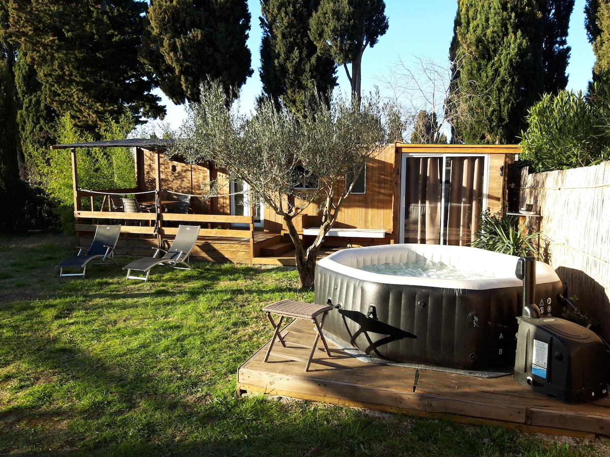 A wooden cottage is visible behind a hot tub situated on a wooden deck. Nearby, loungers and a small table are placed on the grassy area under the shade of trees, creating a serene outdoor space perfect for relaxation.