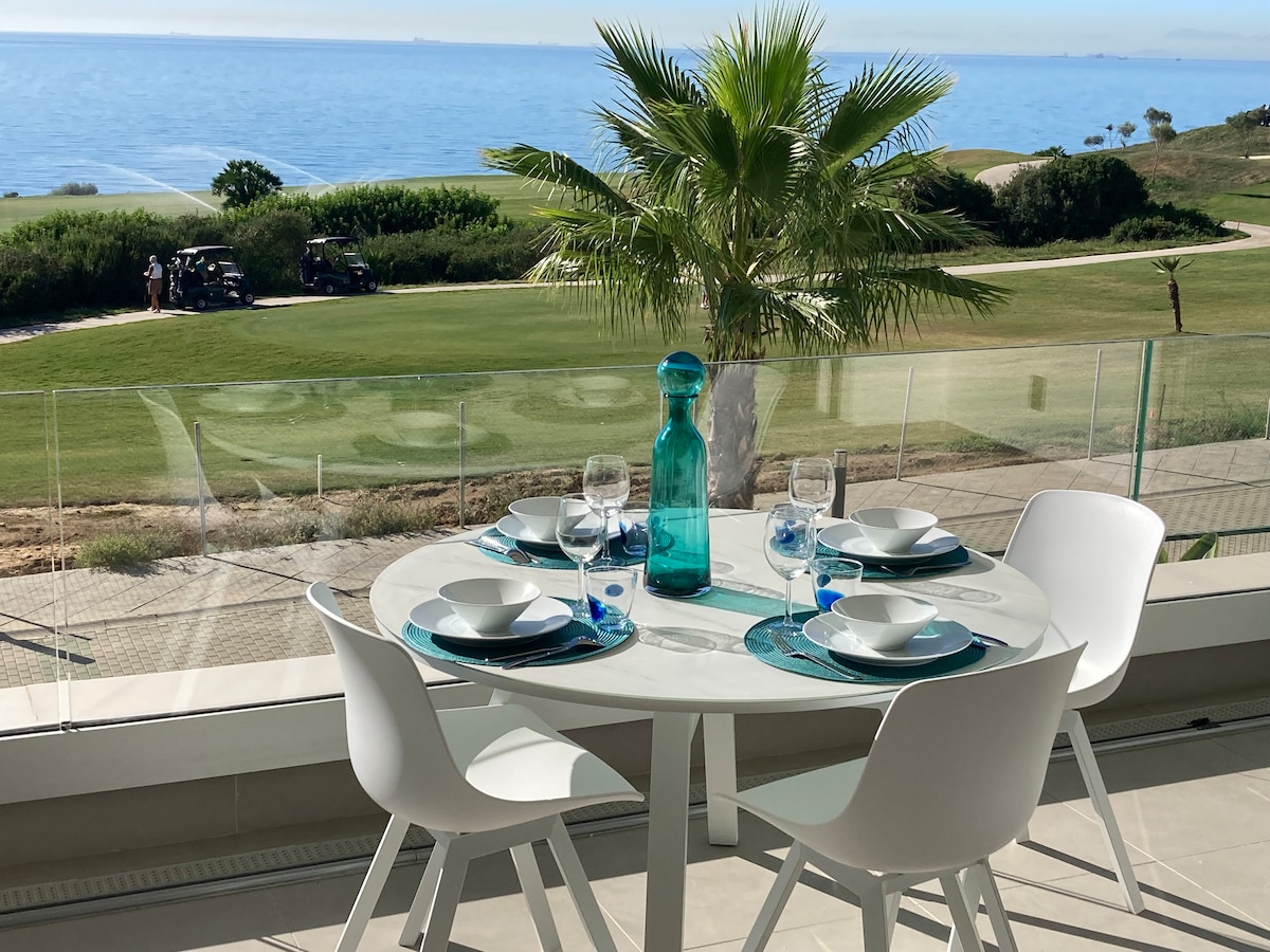 A dining table is set for four, featuring white plates and glasses. A blue water carafe adds a pop of color. The backdrop showcases lush greenery and a scenic view of the sea, enhancing the dining experience with natural beauty.