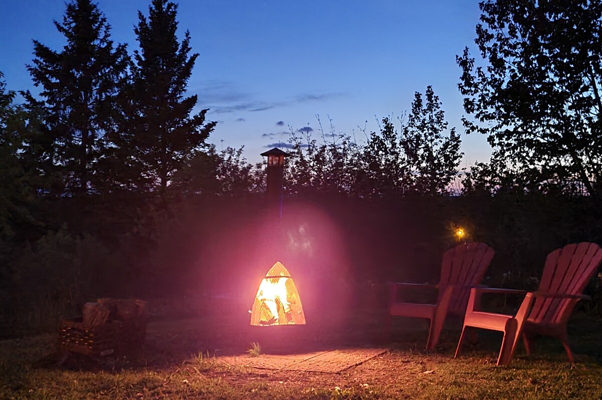 An outdoor fire pit glows warmly against the evening sky, surrounded by green foliage. Two Adirondack chairs are positioned nearby, providing a comfortable seating area. The setting invites relaxation under the stars, with the soft outline of trees framing the scene.