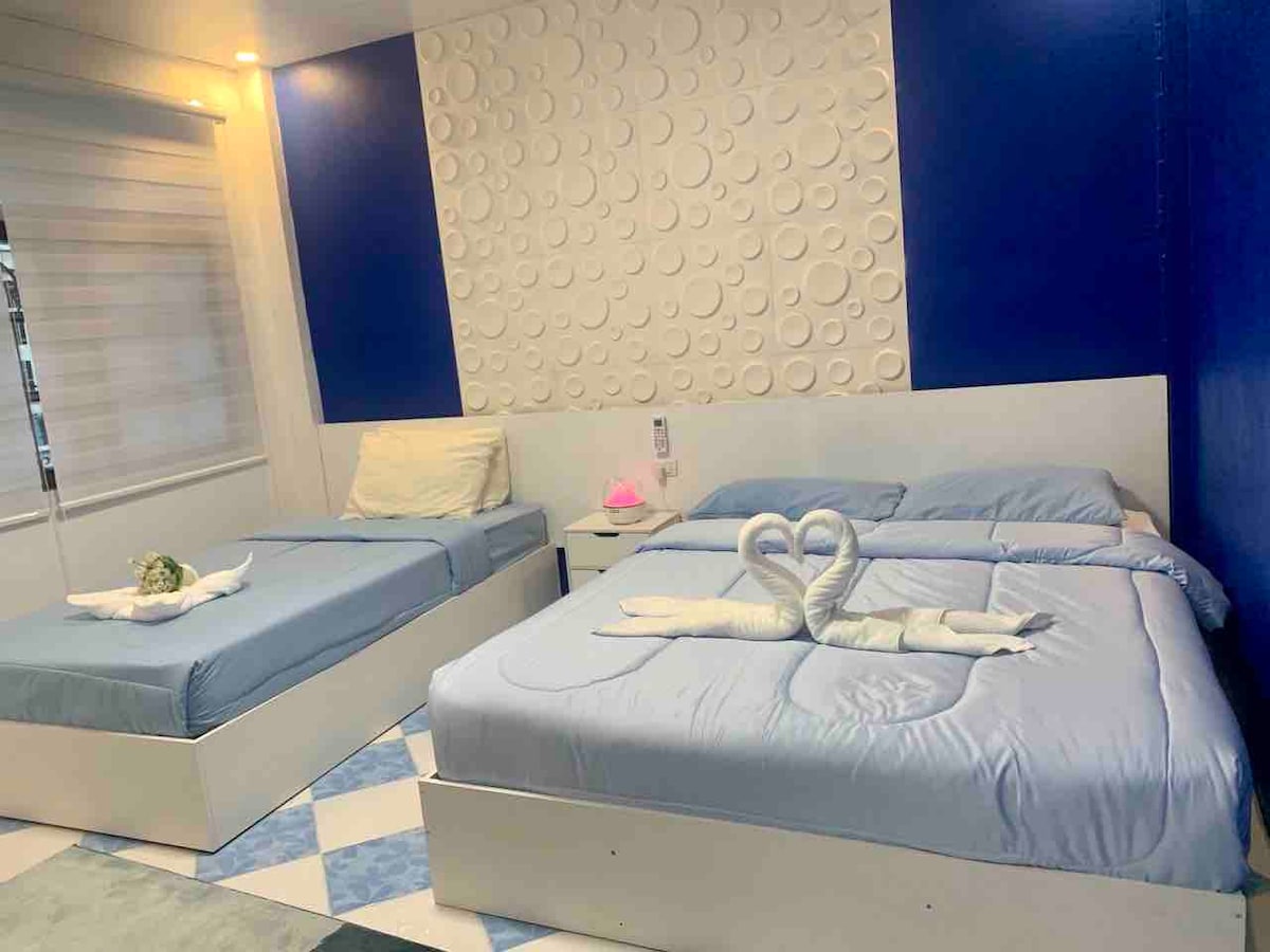 The room contains a queen bed and a twin bed, both adorned with soft blue bedding. A textured white wall contrasts with a vibrant blue feature wall. Decorative towels are arranged in heart shapes on both beds, enhancing the room's welcoming ambiance.