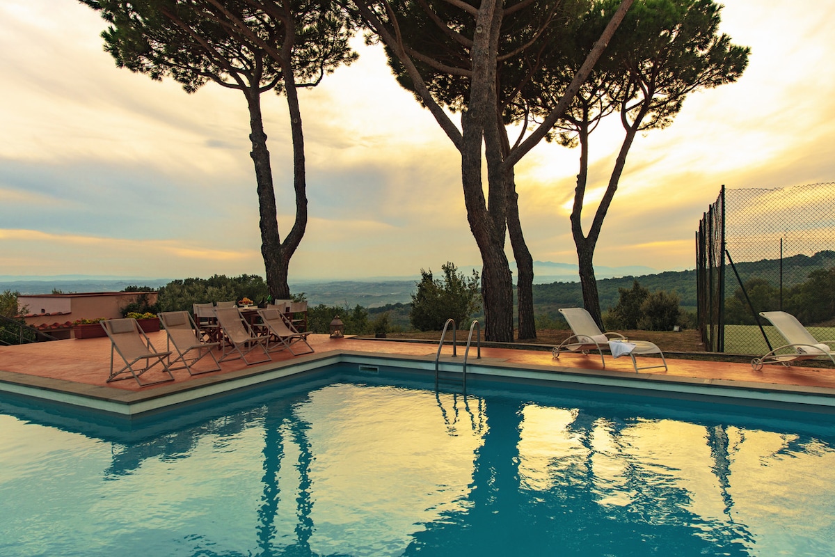 The swimming pool reflects the soft hues of the sunset, bordered by spacious lounge chairs. Tall trees provide shade on one side, while a tennis court is visible in the background. The serene landscape offers expansive views of rolling hills.