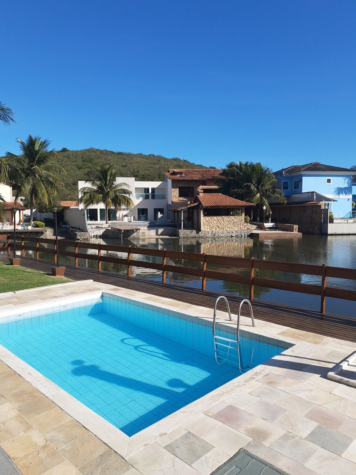 A private pool is framed by a paved deck and features a ladder for easy access. Surrounding palm trees provide shade, while the serene waterway and neighboring houses are visible in the background under a clear blue sky.