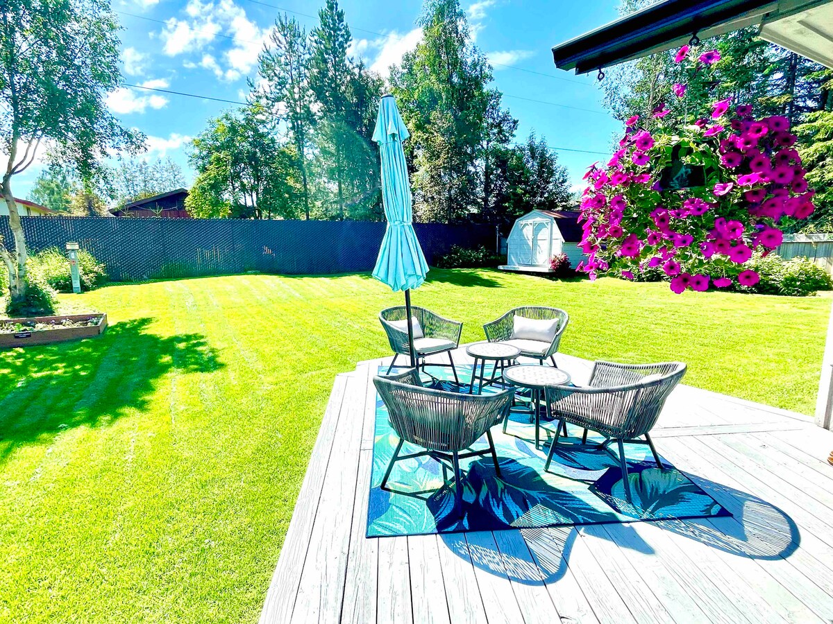 A spacious backyard is presented, featuring a deck with a seating area surrounded by lush green grass. Four chairs are arranged around a table underneath a teal umbrella, while vibrant purple flowers hang nearby, adding color to the tranquil outdoor space.
