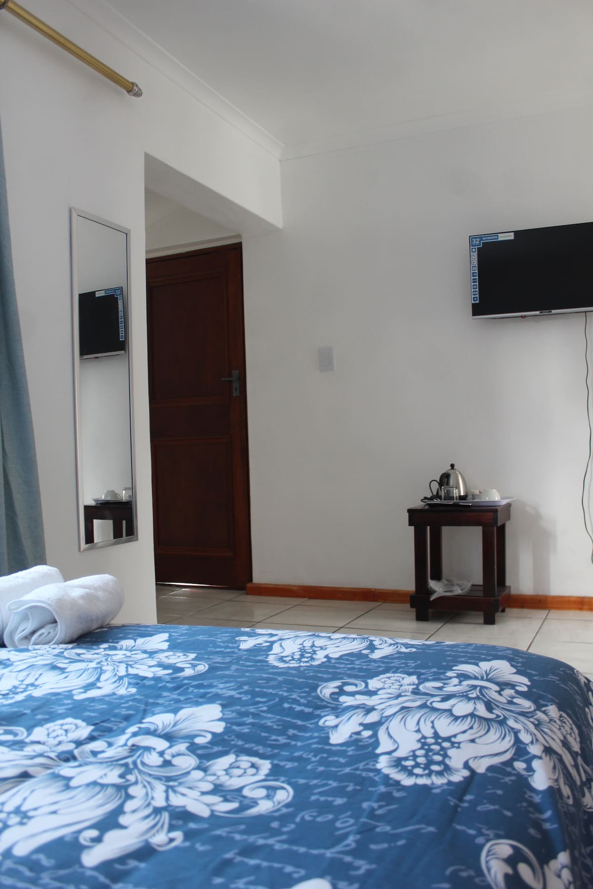 A cozy bedroom space features a spacious bed adorned with a blue and white patterned coverlet. A wall-mounted television is positioned near the door. A mirror and a small table with a kettle and cups are also present, contributing to a functional and comfortable environment.